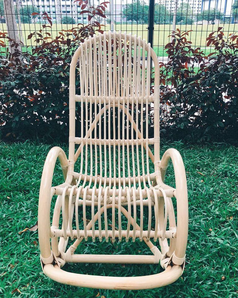 A rattan rocking chair sits on a grassy lawn, with a fence and shrubbery in the background.