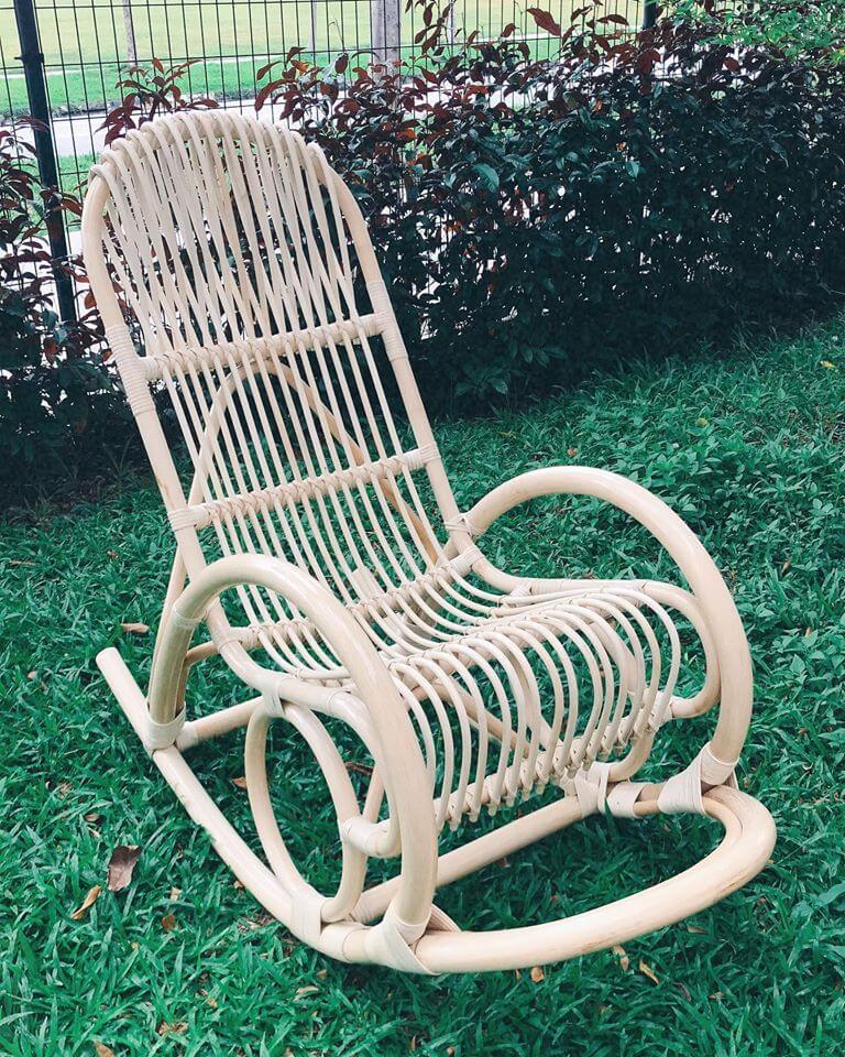 A light-colored wooden rocking chair with a slatted design is placed on a grassy lawn near a hedge and a fence.