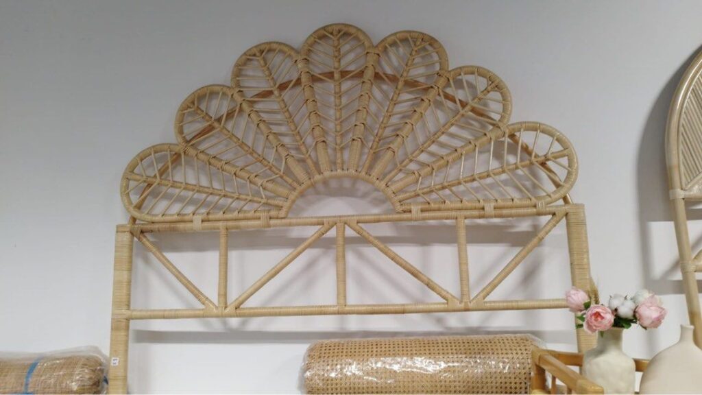 The Floral Natural Rattan Queen Headboard (Clearance), featuring a fan-shaped design, is displayed against a white wall with a small vase of pink and white flowers on the right.