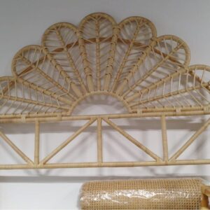 The Floral Natural Rattan Queen Headboard (Clearance), featuring a fan-shaped design, is displayed against a white wall with a small vase of pink and white flowers on the right.