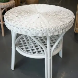 A white wicker table with a round top and open lower shelf is displayed on a gray floor, surrounded by Lounge Chairs and other wicker furniture.