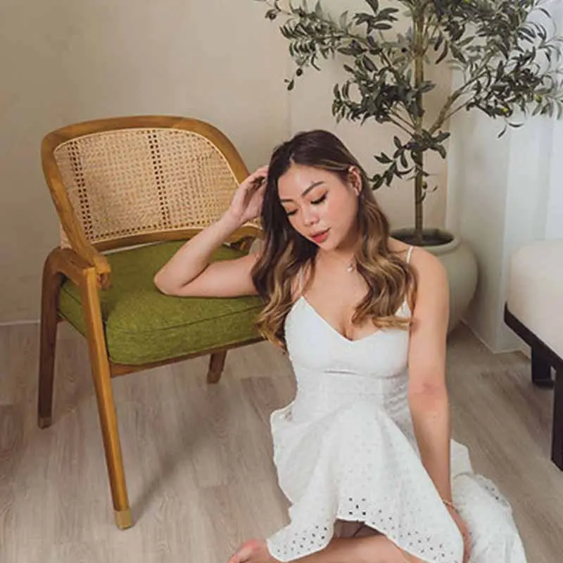 A woman in a white dress sits on a wooden floor next to a wooden chair with a green cushion. An indoor plant is in the background.