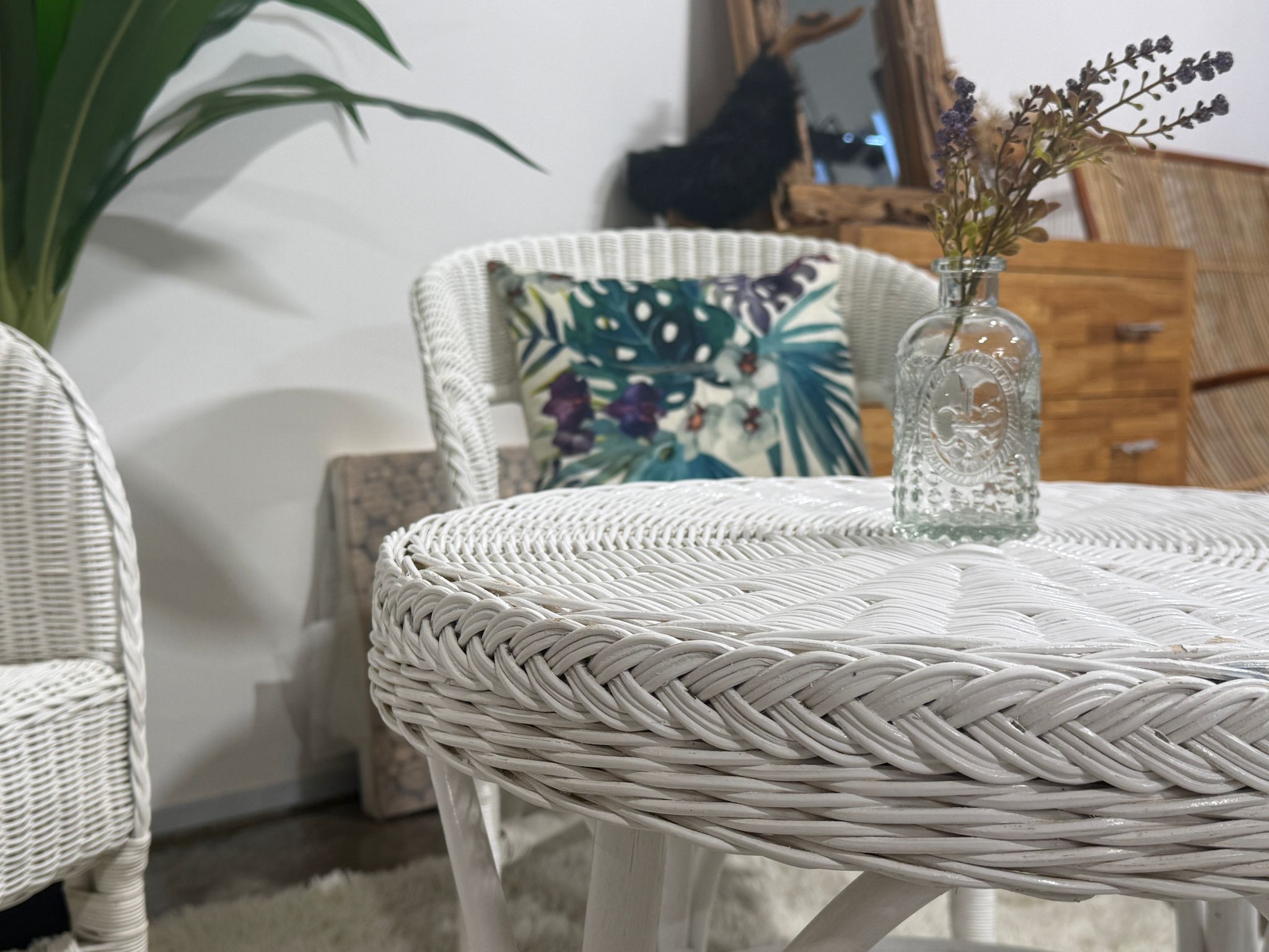 A cozy room with wooden accents features a White Wicker Chair with a tropical print cushion beside another matching chair and a wicker table topped with a small glass vase of dried flowers.