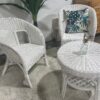 Two White Wicker Chairs and a round table sit on a shaggy rug; one chair features a leaf-patterned cushion, while a small vase with flowers decorates the tabletop.