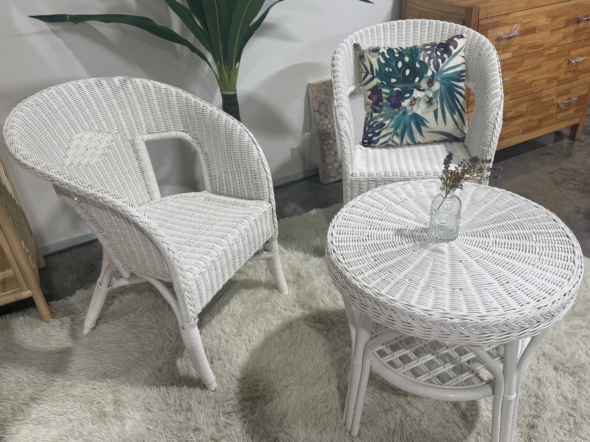 Two White Wicker Chairs and a round table sit on a shaggy rug; one chair features a leaf-patterned cushion, while a small vase with flowers decorates the tabletop.