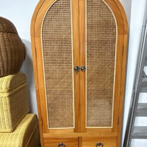 The Perris Arch Cabinet (Clearance Stock) features woven rattan doors, metal handles, and four lower drawers. Wicker baskets and a metal ladder are placed next to it.