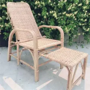 The NN Arm 1 Chair Natural, a woven rattan reclining chair with an extended footrest, is positioned outdoors against a background of lush greenery.