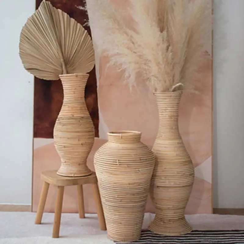 Three woven vases with dried palm leaves and pampas grass are arranged on a stool and the floor against a two-tone background.