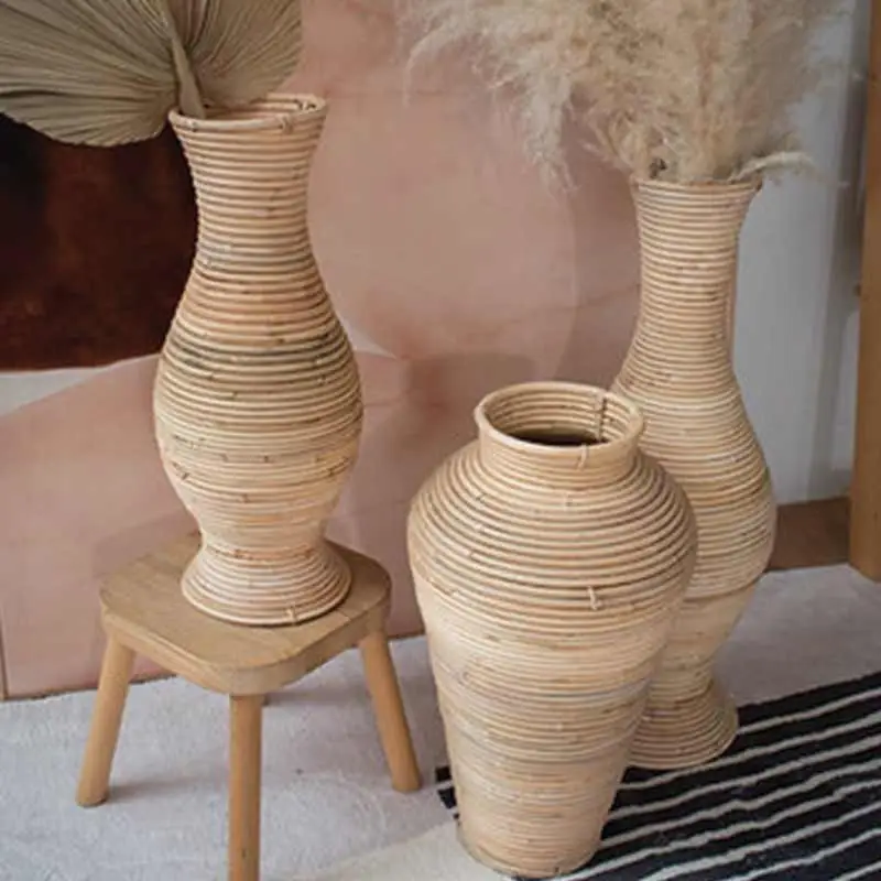 Three tall, woven baskets of varying shapes and sizes are arranged on a wooden stool and the floor, each containing dried foliage. A striped rug and a blurred background are also visible.