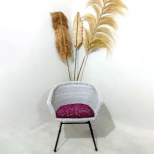 A White Chair with a pink cushion stands on a black metal base in front of a white wall, decorated with tall dried grass arrangements.
