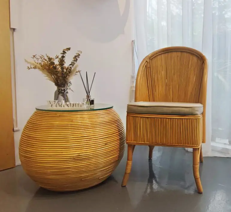 A round rattan table with a glass top and a vase of dried flowers is placed next to the new arrival Zera Chair, a cushioned rattan chair, in a minimalist room with sheer curtains.