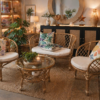 A cozy living room featuring the Bali Rattan Sofa Set (Walnut) with colorful floral cushions, a glass coffee table, lush plants, and warm ambient lighting.
