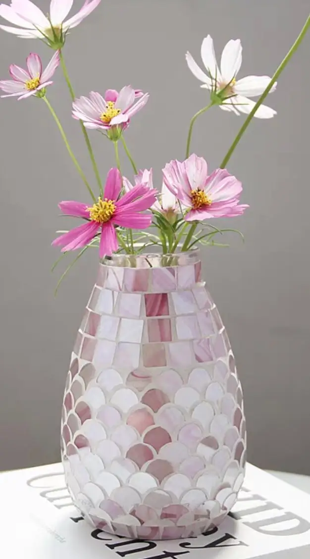 A mosaic-patterned vase holds pink and white cosmos flowers against a plain gray background.