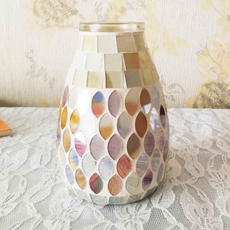 A vase covered with mosaic tiles in various pastel colors sits on a lace tablecloth against a floral wallpaper background.