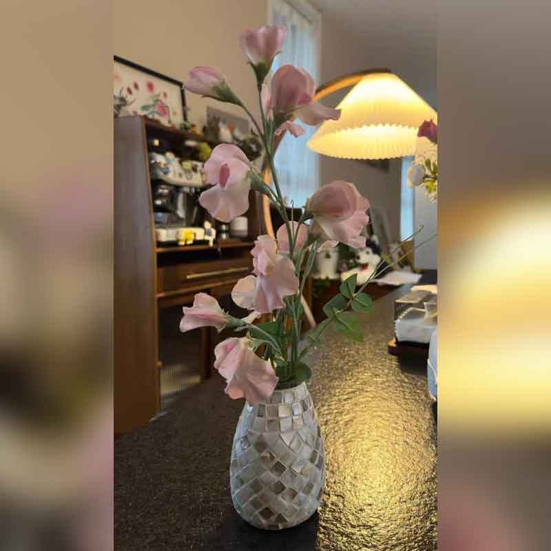 A Creative Mosaic Glass Vase filled with pink flowers is placed on a table next to a lamp, with shelves and wall decor providing the background.