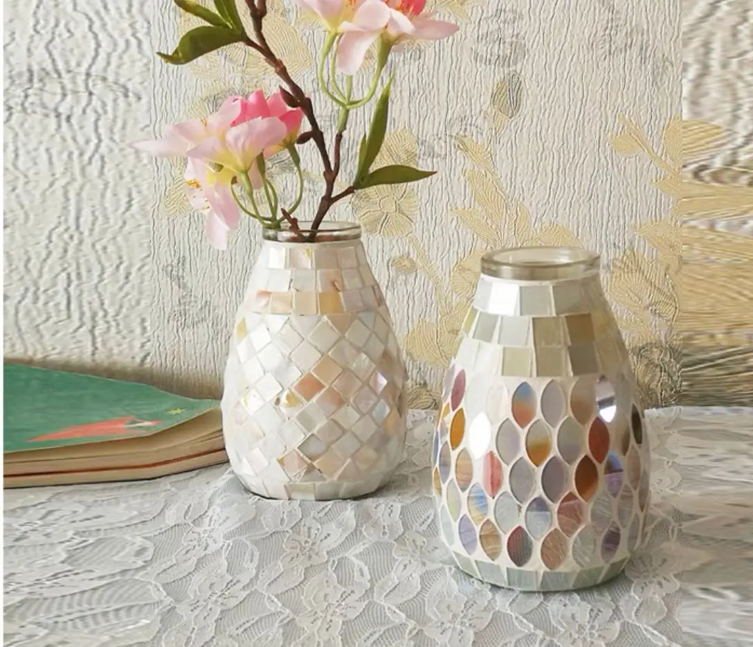 Two Creative Mosaic Glass Vases with pearlescent finishes rest on a lace tablecloth. One vase holds pink flowers, while a closed book sits beside them. In the background, textured wallpaper adds to the scene's elegance.