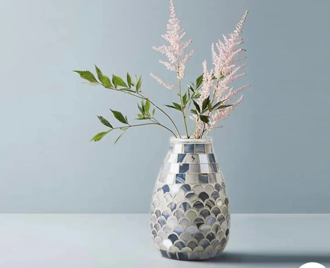 The Creative Mosaic Glass Vase, holding green leaves and pink feathery flowers, is placed on a light-colored surface against a pale blue background.