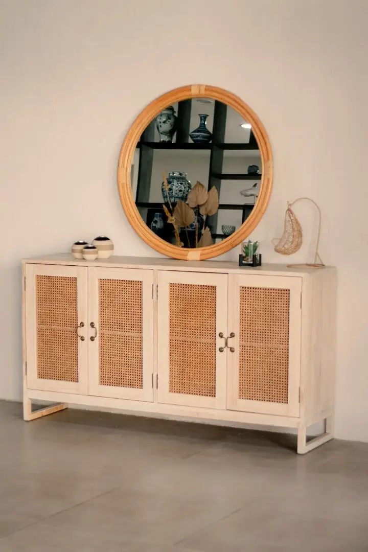 The Pacific Rattan Cabinet, featuring light wood and woven rattan doors, is topped with a round mirror. On the cabinet sit a pair of shoes, a small decorative light, and a sculpture.