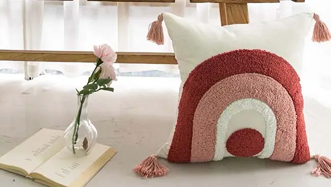 A decorative pillow with a pink and red rainbow pattern rests on the floor beside an open book and a vase holding a pink flower.