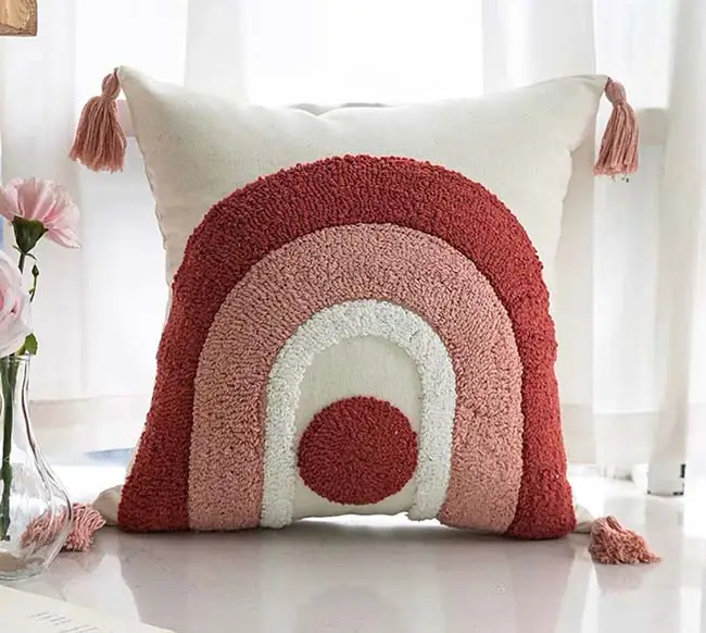 A decorative pillow with a textured rainbow design in shades of red, pink, and white. It features tassels on each corner and is placed on a surface near pink flowers and a window.