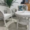 Two white wicker chairs and a round wicker table with a small vase sit on a light rug; a green plant and wooden dresser are in the background.