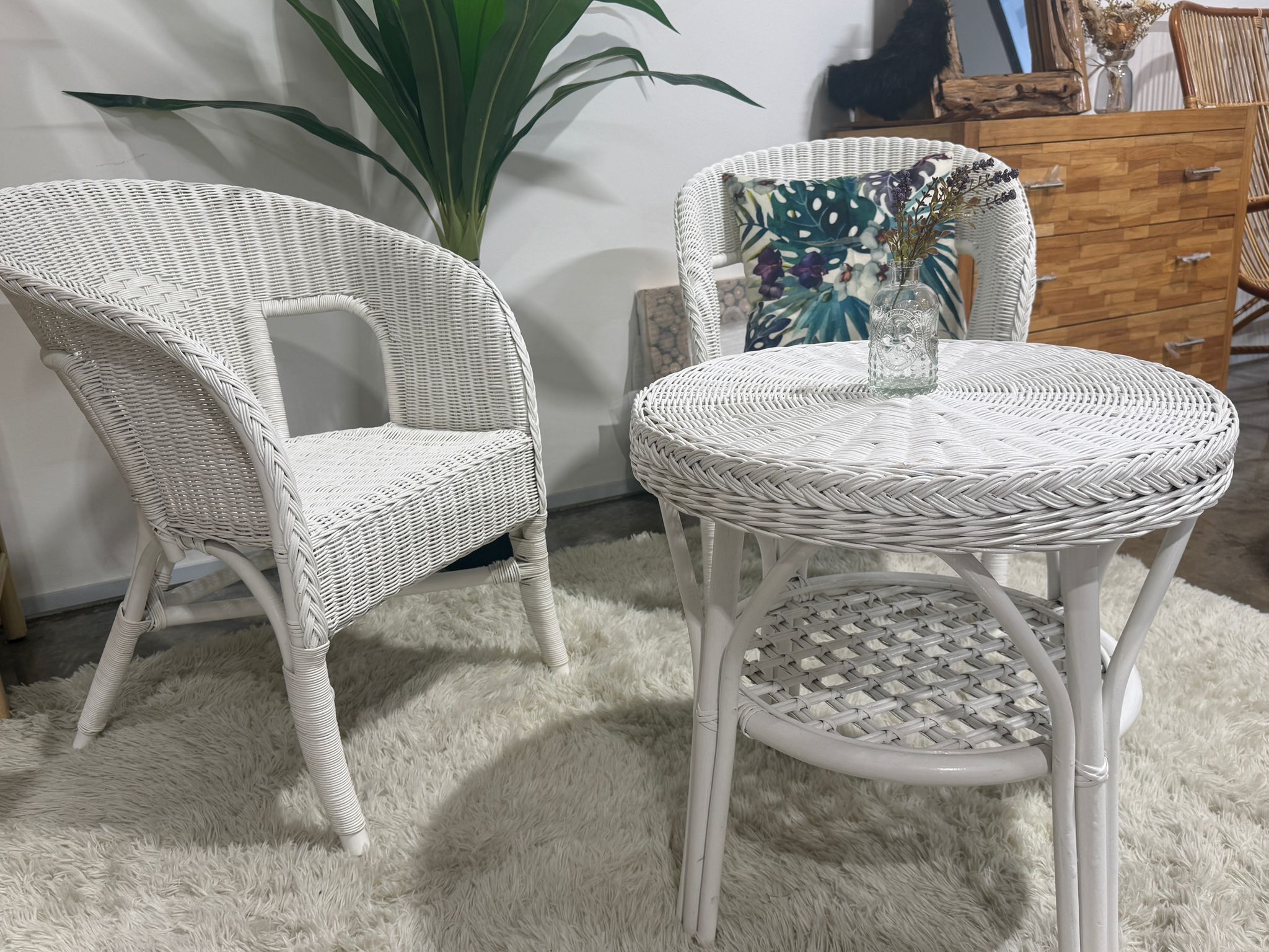 Two white wicker chairs and a round wicker table with a small vase sit on a light rug; a green plant and wooden dresser are in the background.