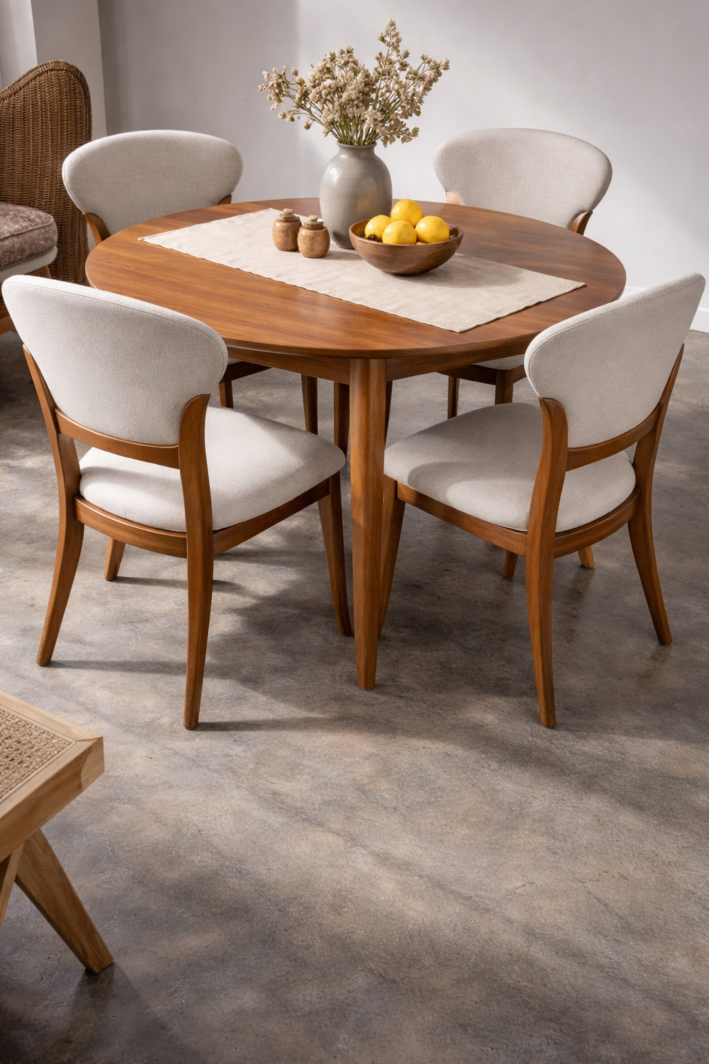 A round wooden dining table with four upholstered chairs, a beige runner, a bowl of lemons, and a vase with dried flowers on top, set on a polished concrete floor. A round wooden dining table with four upholstered chairs, a beige runner, a bowl of lemons, and a vase with dried flowers on top, set on a polished concrete floor.